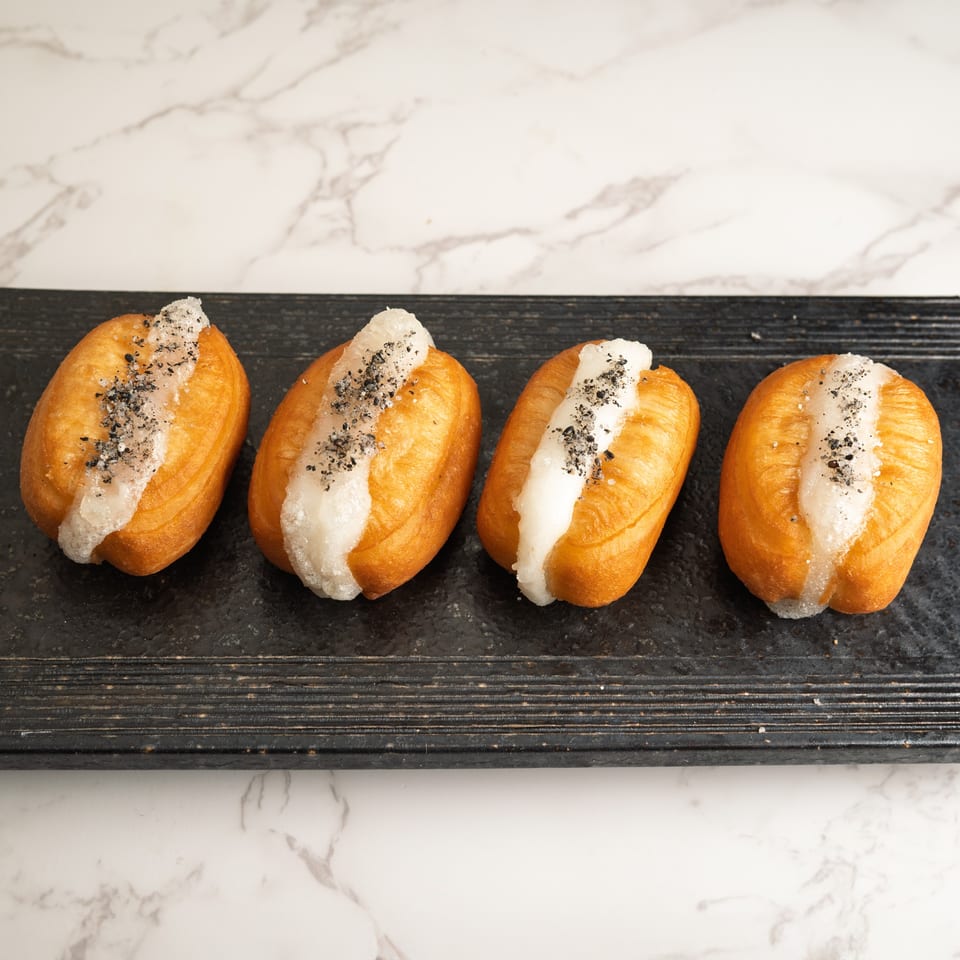 Four golden-brown fried buns with white filling and black sesame seeds on a dark rectangular plate.