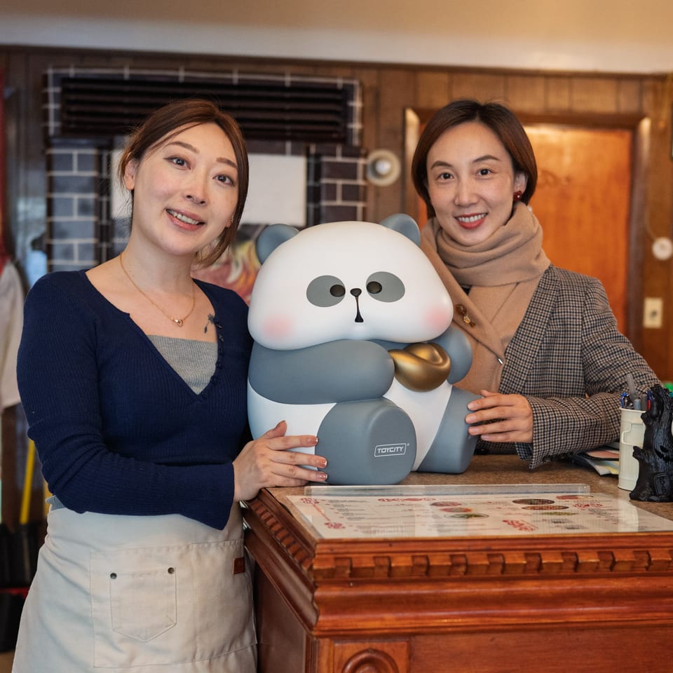 Two women smiling behind a counter with a large panda figurine at Nana Noodle House.