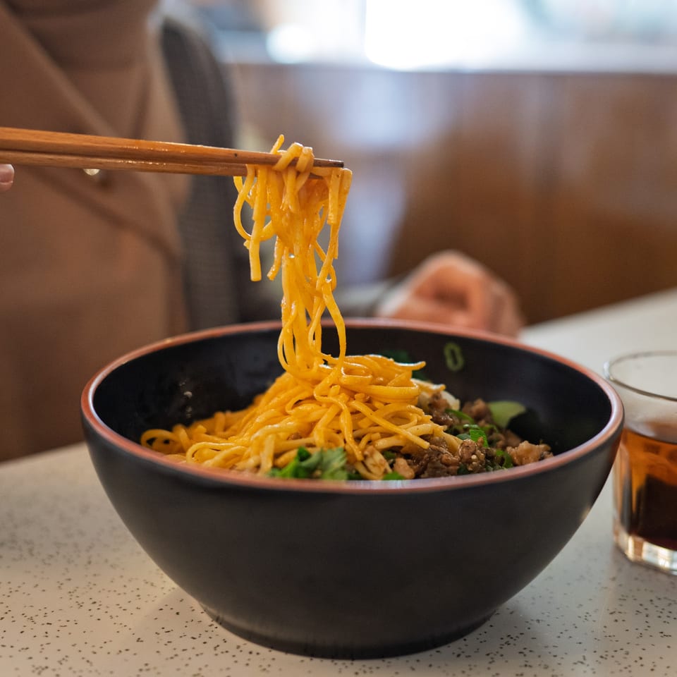 Chopsticks lifting bright yellow noodles from a black bowl of Chinese food with meat and herbs.