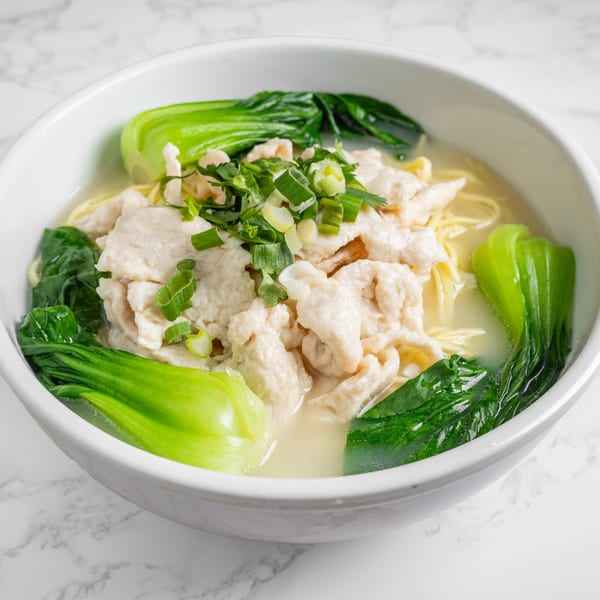 A white bowl of Nana Noodle House chicken noodle soup with bok choy and green onions.