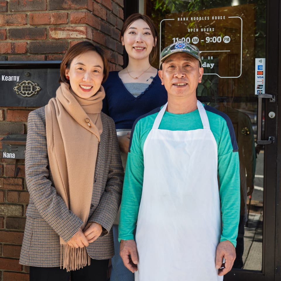 Three smiling people, two women and one man in an apron, stand outside Nana Noodles House.