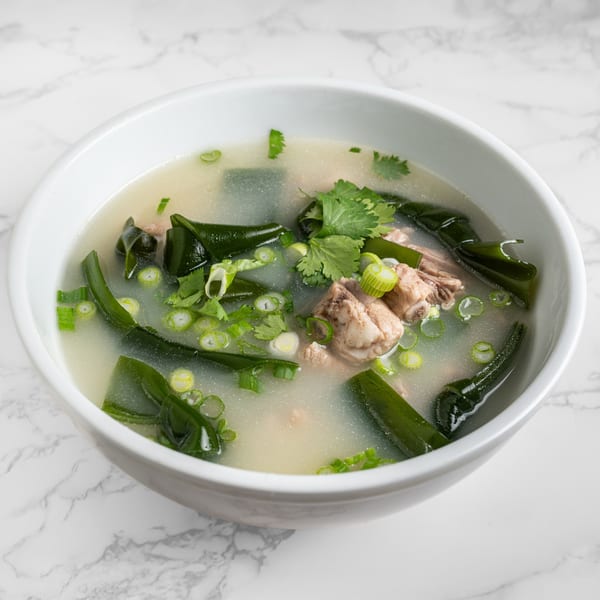 A white bowl of Chinese pork rib soup with seaweed, green onions, and cilantro on marble.