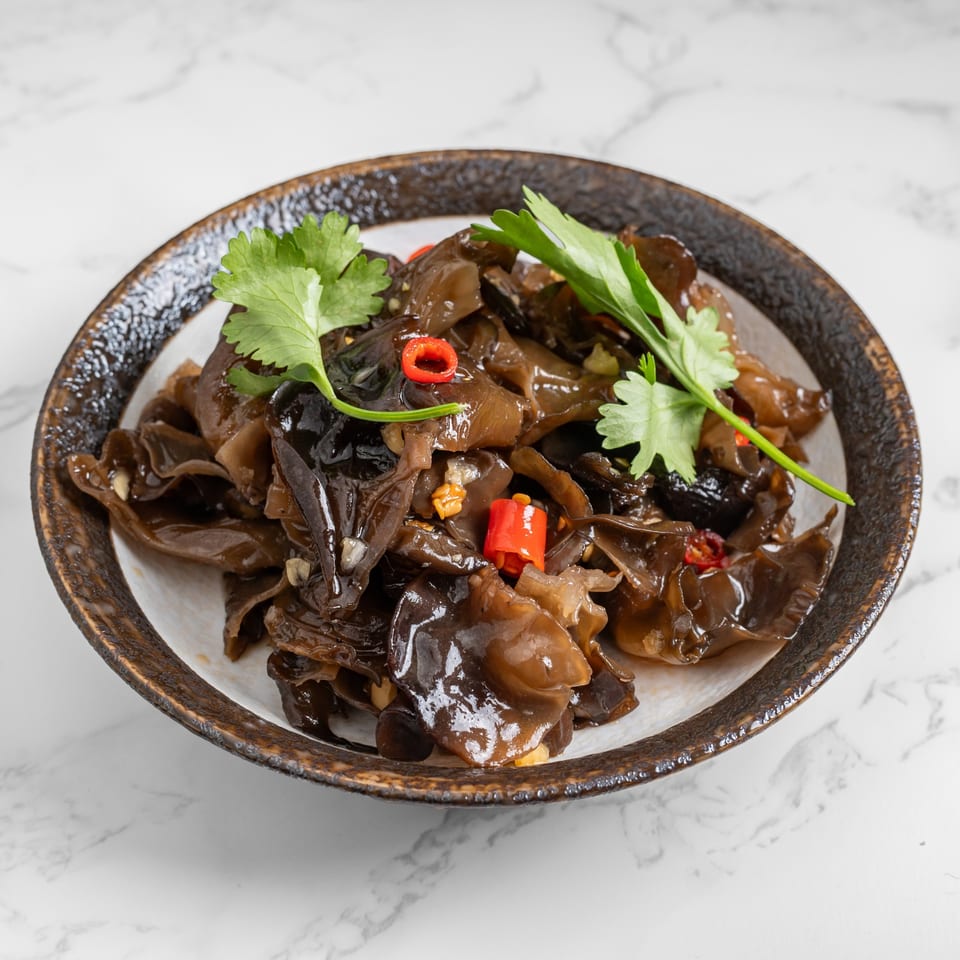 A close-up of a dark brown bowl filled with wood ear mushrooms, red chili, and cilantro.