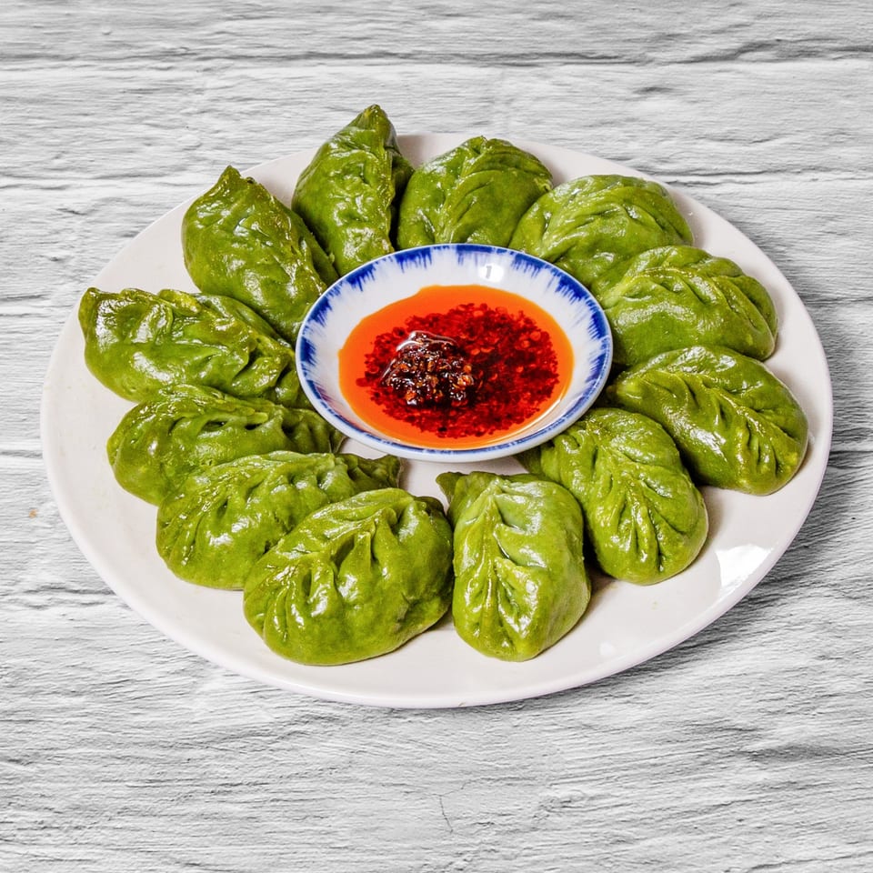 A white plate of green dumplings surrounds a small bowl of chili oil with red flakes.
